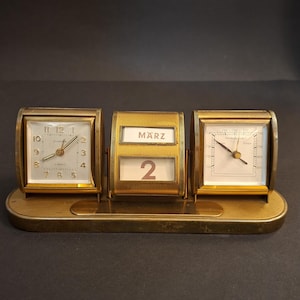 May include: A vintage gold-coloured desk clock with a calendar and barometer. The clock has a square face with Roman numerals. The calendar displays "MARZ 2". The barometer has a white face with a needle. The clock sits on a rectangular base.