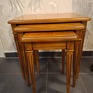 Op de afbeelding: Een set van drie houten nesttafels in verschillende maten. De tafels hebben een warme, bruin-middenbruine afwerking en hebben een rechthoekig blad met een decoratieve rand. De poten zijn taps toelopend en hebben verticale groeven.