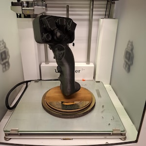 May include: A 3D printed black joystick with multiple buttons and a trigger is being printed on a 3D printer. The printer has a glass build plate and a white base. The joystick is sitting on a wooden base.