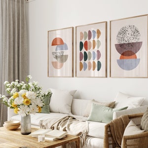 May include: Three abstract art prints with geometric shapes in shades of red, orange, yellow, green, blue, and brown. The prints are framed in natural wood and hung on a white wall above a white couch.