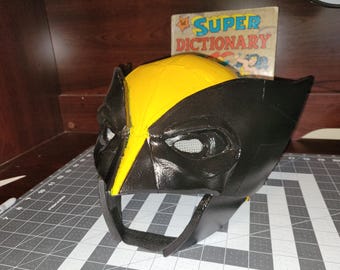 May include: A black and yellow Wolverine mask with a yellow top and black accents. The mask has eye openings with a mesh material and a chin strap. A vintage "Super Dictionary" book is in the background.