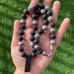 May include: A necklace featuring black lava rock beads and silver-colored metal beads strung on a purple cord. The necklace includes a black rectangular pendant and a silver clasp. The beads are approximately 0.5 inches in diameter.