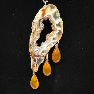 May include: A silver necklace featuring a large, irregularly shaped agate pendant with a hollow center. The agate is primarily beige and white with brown accents. Three teardrop-shaped orange gemstones dangle from the pendant.