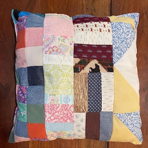 May include: Square patchwork pillow with a variety of fabric patterns and colors. The pillow features a mix of floral, geometric, and solid fabrics in shades of pink, blue, yellow, and green. The pillow is approximately 18 inches square.