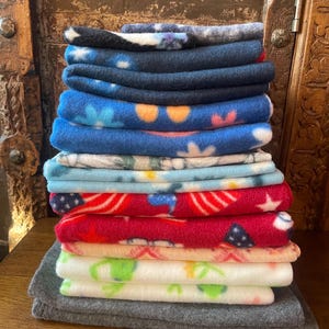 May include: A stack of colorful fleece blankets. The blankets feature various patterns, including stars, flowers, and tie-dye designs. The color palette includes blues, reds, greens, and whites. The blankets are neatly folded and stacked.