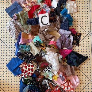 May include: A collection of fabric scraps in various colors and patterns, including floral, polka dots, and geometric designs. A white card with a large black "C" is visible. The fabrics are arranged on a light yellow surface with a grid pattern.