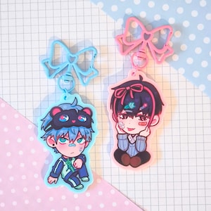 Op de afbeelding: Twee karaktercharms met blauwe en roze strikken. De charms tonen cartoonfiguren met blauw en zwart haar. Een personage draagt een blauw en groen trainingspak en een blauw oogmasker. Het andere personage draagt een blauwe trui en heeft roze haar.