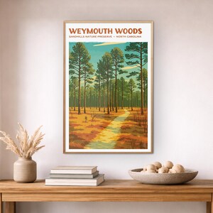 May include: A framed art print of Weymouth Woods, a North Carolina nature preserve. The artwork shows a path through a forest of tall pine trees under a blue sky. The print is displayed on a wooden console table with decorative items.
