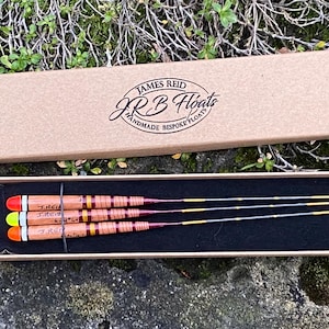 May include: Three wooden fishing floats in a brown box with a black interior. The box has a label that reads "James Reid J.R.B Floats Handmade Bespoke Floats". The floats are red, yellow, and orange with black and gold accents.