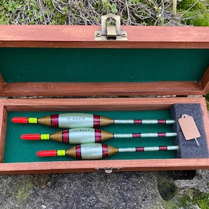 May include: Three wooden fishing floats with red, yellow, and white stripes and the name "J. Reed" written on them. The floats are in a wooden box with a green felt lining.