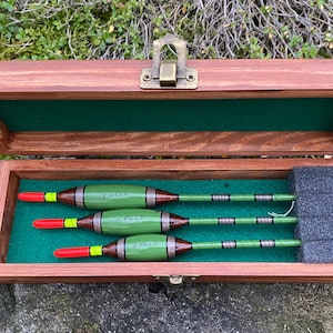 Könnte beinhalten: Eine Holzkiste mit grüner Filz-Auskleidung und drei grünen und braunen Angelbojen mit schwarzen und weißen Streifen. Die Bojen sind mit "3 REED", "2 REED" und "1 REED" beschriftet.