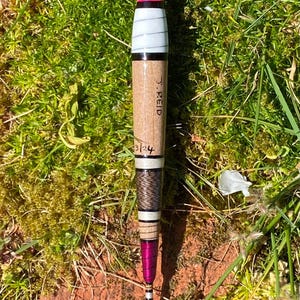 Pode incluir: Uma bóia de pesca com um topo rosa brilhante, listras brancas e pretas e um corpo de madeira. A bóia tem o nome "J. REID" e os números "3/24". A base é de cor roxa escura.