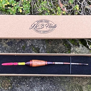 Könnte beinhalten: Ein hölzerner Angel-Schwimmer mit roten, gelben und weißen Akzenten in einer braunen Box. Die Box trägt den Text "James Reid JR B Floats Handmade Bespoke Floats" auf der Oberseite.
