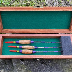 Peut inclure: Trois flotteurs de pêche en bois avec des accents violets, jaunes et rouges sont présentés dans une boîte en bois avec un revêtement en feutre vert.