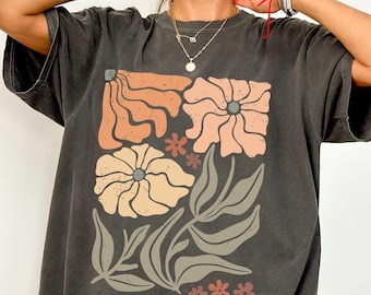 May include: Dark gray t-shirt featuring a floral design in shades of peach, tan, and brown. The graphic includes stylized flowers and leaves. The shirt has a relaxed fit and short sleeves, suitable for casual wear.