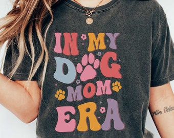 May include: A dark gray t-shirt with the text "In My Dog Mom Era" in colorful retro-style lettering. The shirt features paw prints and stars.