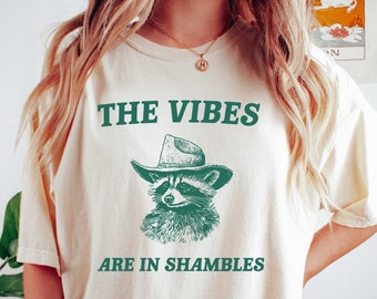 May include: A cream colored t-shirt with a green raccoon wearing a cowboy hat and the text "THE VIBES ARE IN SHAMBLES". The shirt color is ivory.