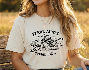May include: Cream-colored t-shirt with the text "FERAL AUNTS SOCIAL CLUB" and a graphic of a person riding a tiger. The person is wearing a hat and holding a rope. The shirt is worn by a person sitting on a wooden bench.