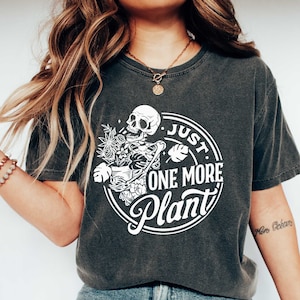 Może przedstawiać: Szary t-shirt z białym nadrukiem przedstawiającym szkielet trzymający doniczkę z roślinami. Nadruk głosi "Just One More Plant".