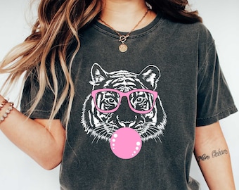May include: A dark gray t-shirt with a white graphic of a tiger wearing pink glasses and blowing a pink bubble. The shirt color is "pepper".