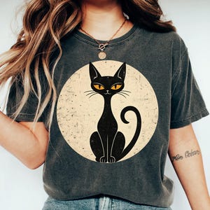 May include: A charcoal gray short-sleeve t-shirt featuring a large circular graphic on the chest. The graphic depicts a stylized black cat with bright orange eyes centered within a distressed, cream-colored circle. The shirt has a crew neckline.