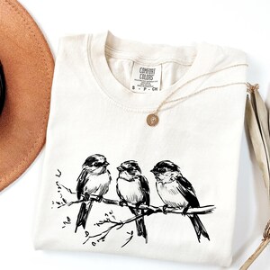 May include: Cream-colored t-shirt featuring a black and white illustration of three birds perched on a branch. The shirt has a crew neck and short sleeves. A gold necklace with a pendant is draped on the shirt. A brown hat and dried plant are in the background.