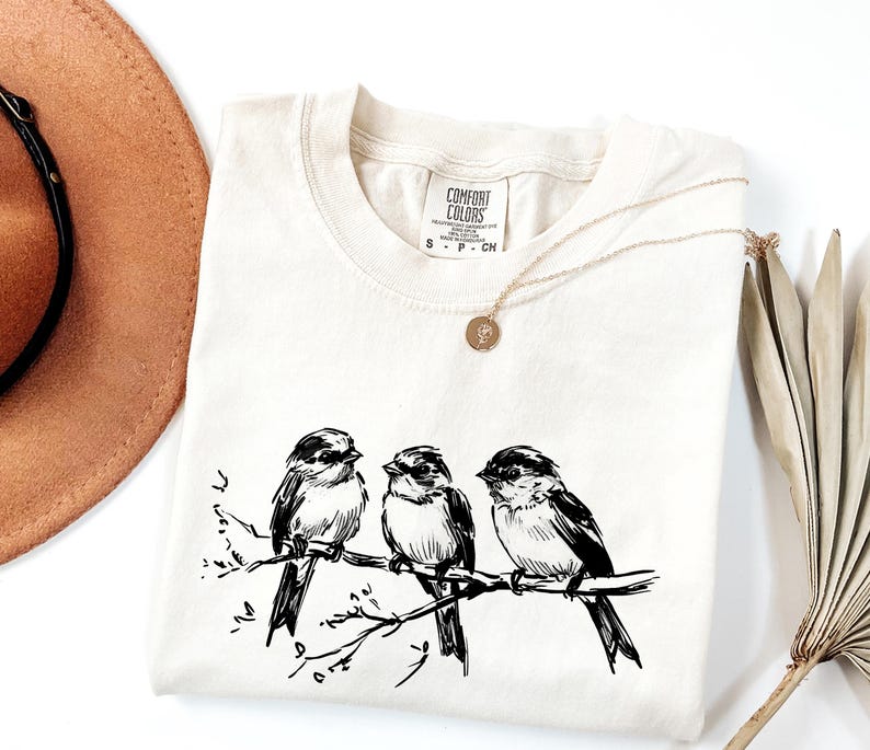 May include: Cream-colored t-shirt featuring a black and white illustration of three birds perched on a branch. The shirt has a crew neck and short sleeves. A gold necklace with a pendant is draped on the shirt. A brown hat and dried plant are in the background.