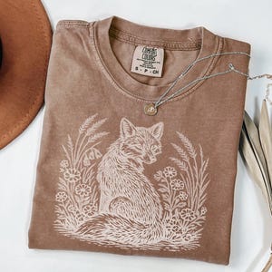 Vintage-style espresso color t-shirt with a hand-drawn white fox and botanical nature scene. The shirt has a "lived-in" washed look typical of Comfort Colors apparel. The fox is looking back over its shoulder, framed by delicate floral and wheat accents, creating a cottagecore or forest-inspired aesthetic.