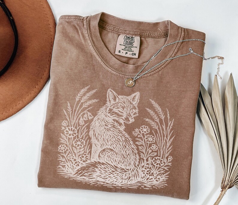 Vintage-style espresso color t-shirt with a hand-drawn white fox and botanical nature scene. The shirt has a "lived-in" washed look typical of Comfort Colors apparel. The fox is looking back over its shoulder, framed by delicate floral and wheat accents, creating a cottagecore or forest-inspired aesthetic.