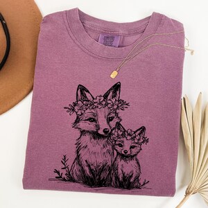 May include: A dusty rose Comfort Colors t-shirt featuring a black ink illustration of two foxes wearing floral crowns. The foxes are drawn in a detailed, artistic style. A gold necklace is draped across the shirt.