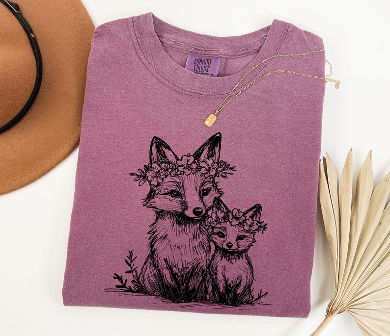 May include: A dusty rose Comfort Colors t-shirt featuring a black ink illustration of two foxes wearing floral crowns. The foxes are drawn in a detailed, artistic style. A gold necklace is draped across the shirt.