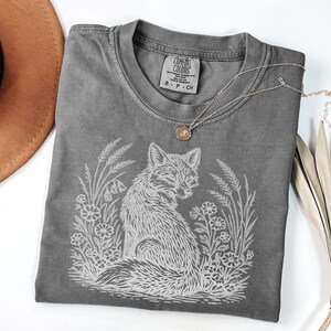 Pepper color Comfort Colors t-shirt with a white graphic of a fox sitting in a field of flowers and tall grass, styled with rustic accessories on a white background.