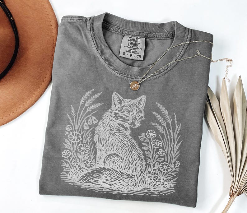 Pepper color Comfort Colors t-shirt with a white graphic of a fox sitting in a field of flowers and tall grass, styled with rustic accessories on a white background.
