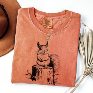May include: A coral-colored t-shirt featuring a black and white illustration of a squirrel sitting on a tree stump. The shirt has a crew neck and a small tag on the collar. A gold necklace with a pendant is draped across the shirt.