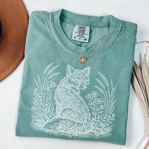 May include: A teal Comfort Colors t-shirt featuring a white illustration of a fox surrounded by flowers and foliage. A silver necklace with a small pendant rests on the shirt. A brown hat and dried plant are in the background.