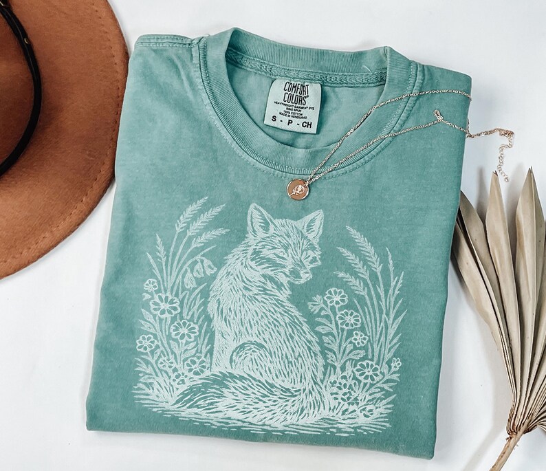 May include: A teal Comfort Colors t-shirt featuring a white illustration of a fox surrounded by flowers and foliage. A silver necklace with a small pendant rests on the shirt. A brown hat and dried plant are in the background.