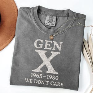 May include: A gray Comfort Colors t-shirt with the text "GEN X 1965-1980 WE DON'T CARE" in white. A gold necklace with a pendant rests on the shirt. A brown hat is visible in the upper left corner.