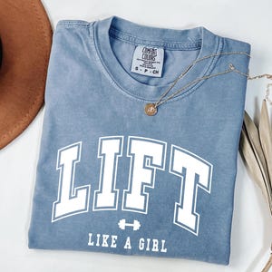 Pode incluir: Uma camiseta azul com a palavra "LIFT" em letras grandes brancas e a frase "LIKE A GIRL" abaixo. Um pequeno gr&aacute;fico de halteres est&aacute; entre as palavras. Um colar de ouro com um pingente est&aacute; na camiseta.