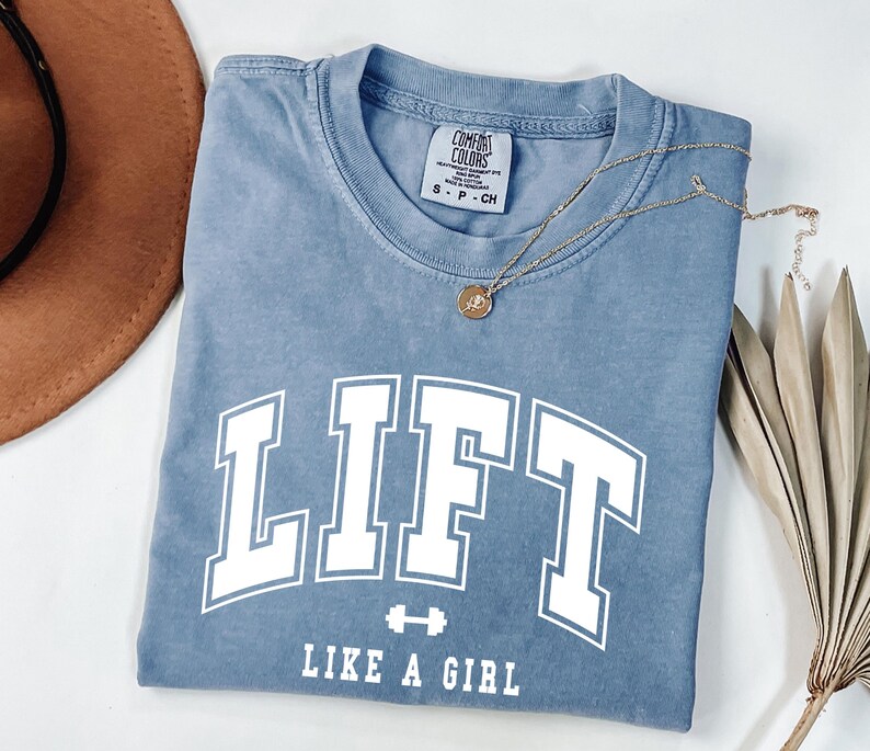 Pode incluir: Uma camiseta azul com a palavra "LIFT" em letras grandes brancas e a frase "LIKE A GIRL" abaixo. Um pequeno gr&aacute;fico de halteres est&aacute; entre as palavras. Um colar de ouro com um pingente est&aacute; na camiseta.