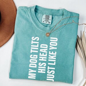 以下が含まれることがあります： 「MY DOG TILTS HIS HEAD JUST LIKE YOU」の白い文字が入った、ターコイズ色のComfort Colors Tシャツ。ペンダント付きのゴールドネックレスと茶色の帽子も写っています。