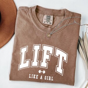 Pode incluir: Uma camiseta marrom com a palavra "LIFT" em letras brancas grandes e a frase "LIKE A GIRL" abaixo. Um pequeno gr&aacute;fico de halteres est&aacute; entre as palavras. Um colar de prata com um pequeno pingente est&aacute; na camisa.