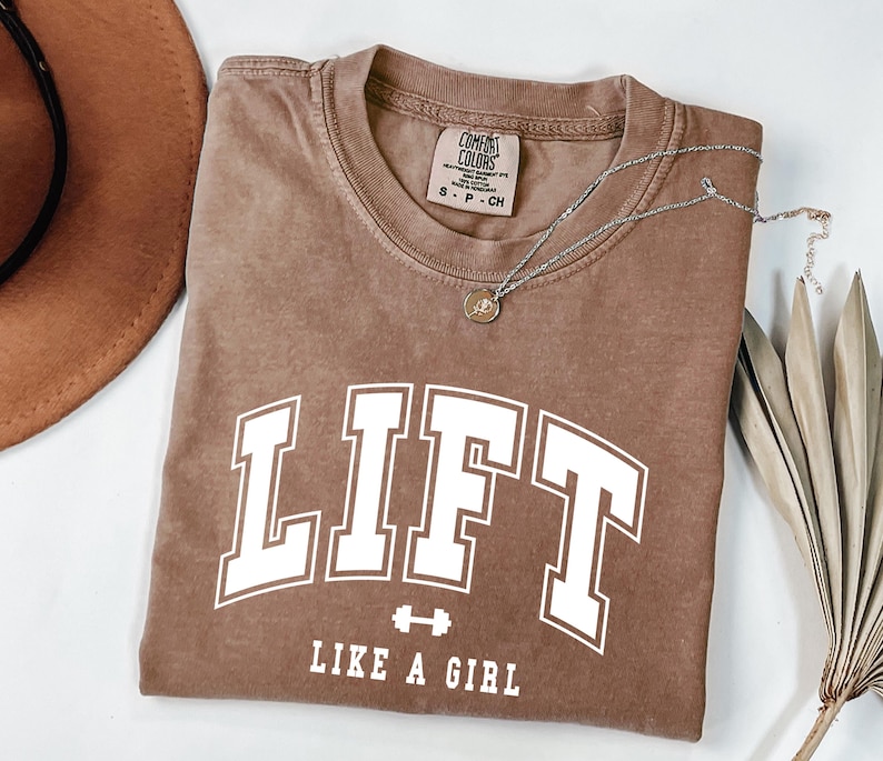Pode incluir: Uma camiseta marrom com a palavra "LIFT" em letras brancas grandes e a frase "LIKE A GIRL" abaixo. Um pequeno gr&aacute;fico de halteres est&aacute; entre as palavras. Um colar de prata com um pequeno pingente est&aacute; na camisa.