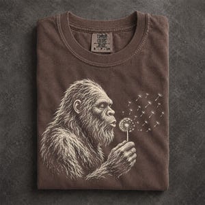 May include: A brown crew neck t-shirt featuring a white graphic illustration of a hairy ape-like creature blowing on a dandelion, with the Comfort Colors brand label visible at the inner neckline showing size S-P-CH and garment details.