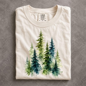 May include: A cream-colored short-sleeve t-shirt featuring a watercolor-style graphic of a dense forest with various shades of green and blue pine trees. The inner neck label displays the text Comfort Colors with size S CH printed below.