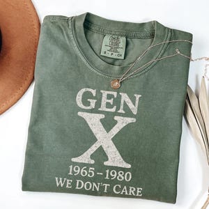 May include: An olive green t-shirt with the text "GEN X 1965-1980 WE DON'T CARE" in white. A gold necklace with a pendant is draped across the shirt. A brown hat and dried plant are in the background.