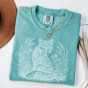 May include: A teal Comfort Colors t-shirt featuring a white graphic of a fox surrounded by flowers and foliage. A gold necklace with a small pendant rests on the shirt. The shirt is size S-P-CH.