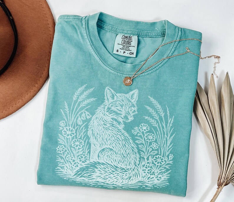 May include: A teal Comfort Colors t-shirt featuring a white graphic of a fox surrounded by flowers and foliage. A gold necklace with a small pendant rests on the shirt. The shirt is size S-P-CH.