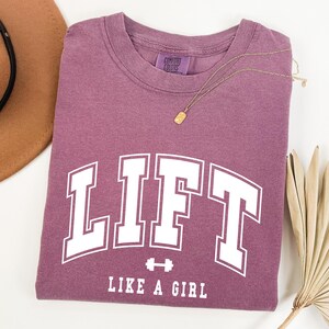 Pode incluir: Uma camiseta de cor malva com a palavra "LIFT" em letras brancas grandes, com "LIKE A GIRL" abaixo. Um colar de ouro e um chap&eacute;u marrom tamb&eacute;m est&atilde;o na imagem. A camiseta &eacute; feita de um material macio.