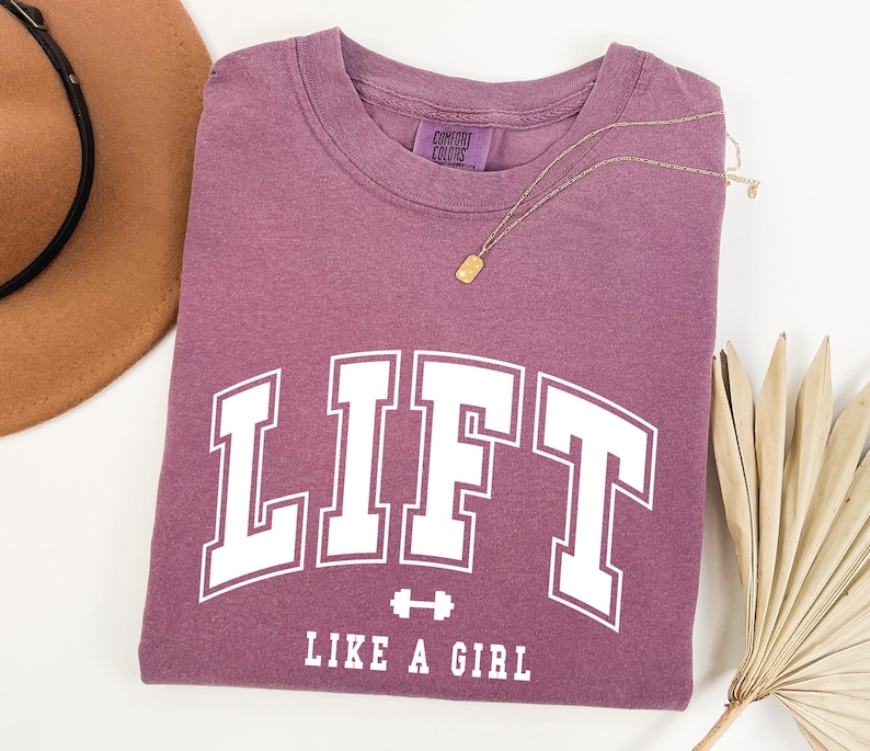 Pode incluir: Uma camiseta de cor malva com a palavra "LIFT" em letras brancas grandes, com "LIKE A GIRL" abaixo. Um colar de ouro e um chap&eacute;u marrom tamb&eacute;m est&atilde;o na imagem. A camiseta &eacute; feita de um material macio.