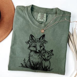 May include: An olive green Comfort Colors t-shirt featuring a black illustration of two foxes wearing floral crowns. A gold necklace with a small pendant rests on the shirt. The shirt is folded, with a brown hat visible in the background.
